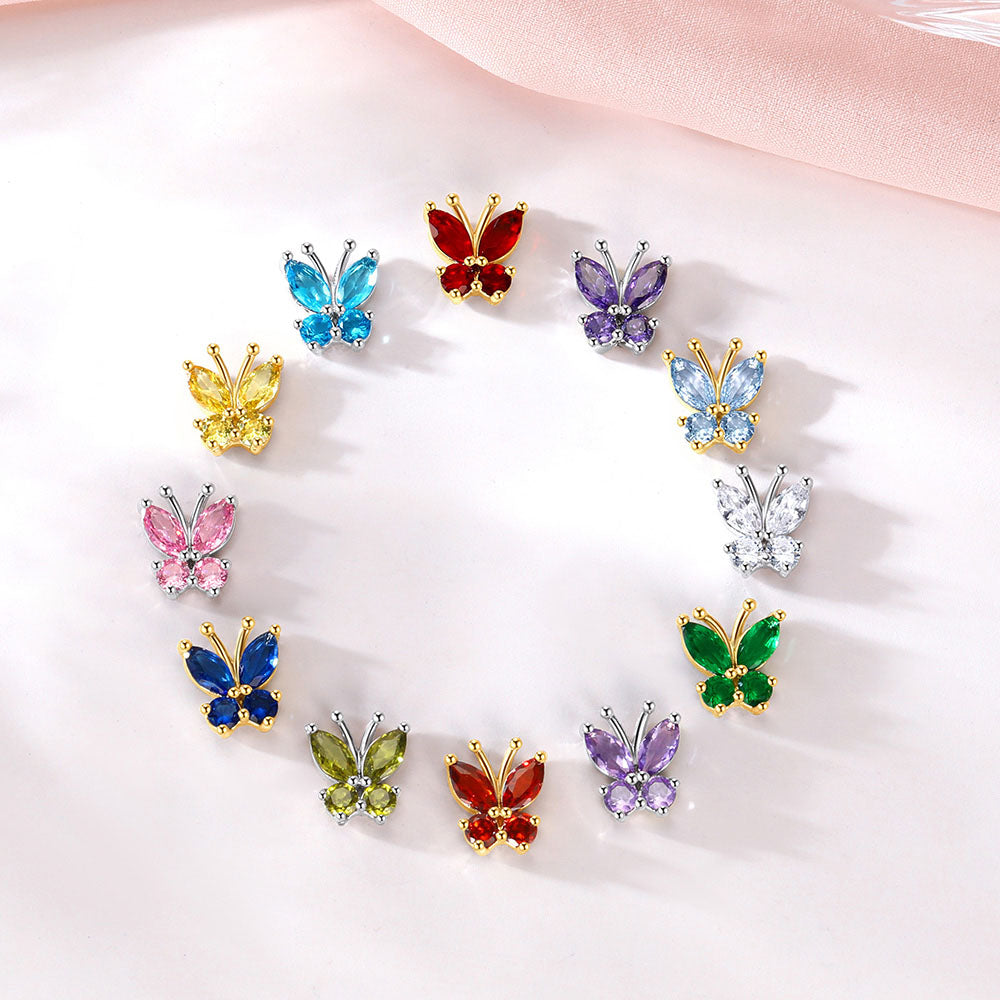 Twelve colorful butterfly stud earrings pieces arranged in a circle on a soft white surface with a pale pink fabric in the background. The butterflies feature gold-tone metal settings and various colored stones including blue, red, purple, yellow, pink, green, and silver.