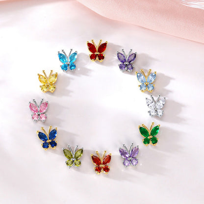 Twelve colorful butterfly stud earrings pieces arranged in a circle on a soft white surface with a pale pink fabric in the background. The butterflies feature gold-tone metal settings and various colored stones including blue, red, purple, yellow, pink, green, and silver.