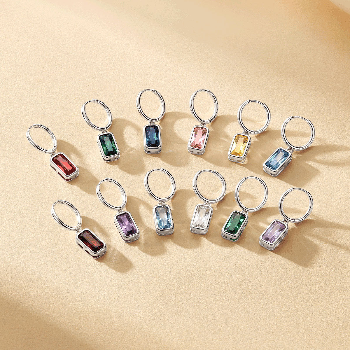Twelve silver birthstone earrings with rectangular bezel-set gemstones in various colors, arranged in two rows on a warm beige textured background.