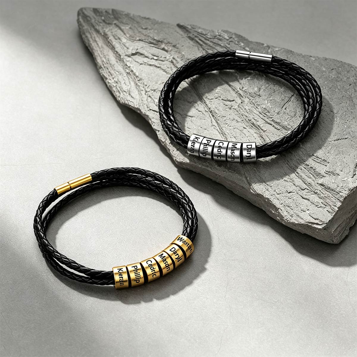 Two black braided cord bracelets with numeric lock combination charms are displayed on a light gray surface, one resting on a textured gray stone slab and the other below it. One bracelet features gold-toned combination dials, and the other features silver-toned combination dials.