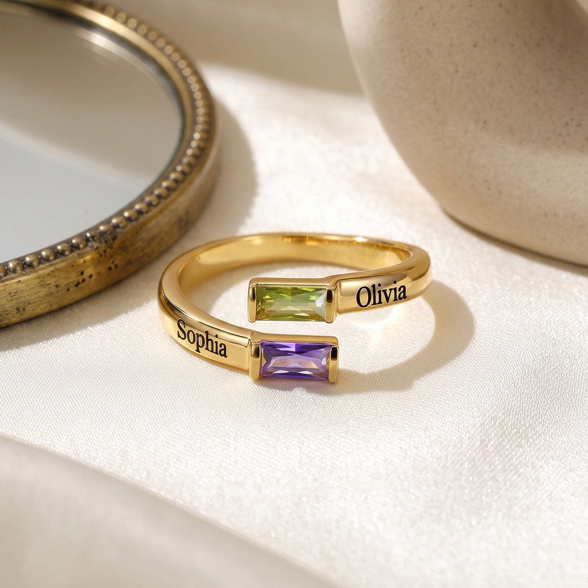 Two gold custom birthstone rings engraved with 'Sophia' and 'Olivia', featuring rectangular baguette-cut gemstones in green and purple, displayed on a cream fabric surface.