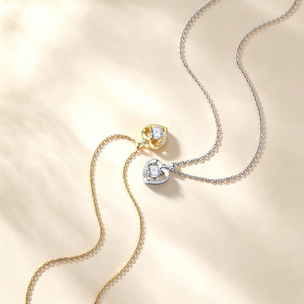 Two heart-shaped pendant necklaces on delicate chains, one in yellow gold and one in silver, both featuring clear heart-shaped gemstones, displayed on a warm beige textured background.