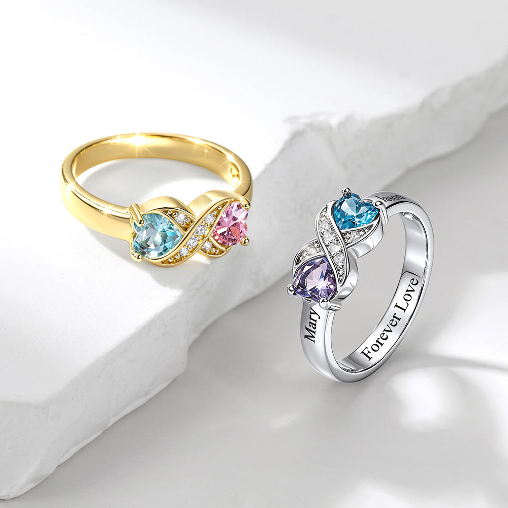 Two infinity rings displayed on a white surface with a light gray stone prop. One ring is yellow gold with light blue and pink heart-shaped gemstones and small clear accent stones.