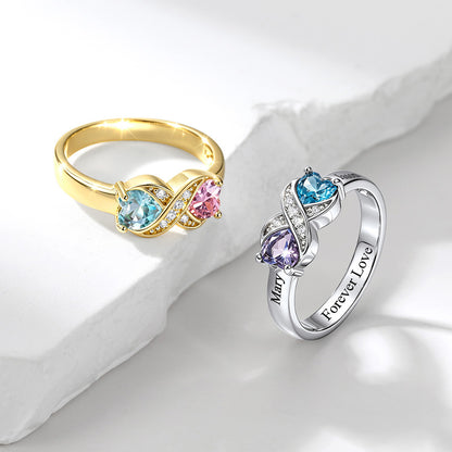 Two infinity rings displayed on a white surface with a light gray stone prop. One ring is yellow gold with light blue and pink heart-shaped gemstones and small clear accent stones.