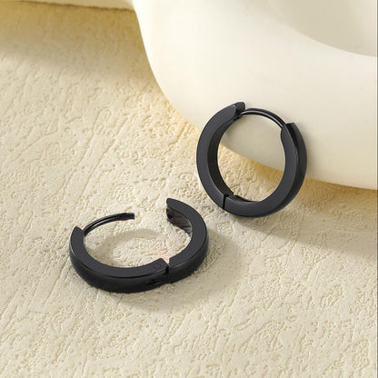 Two matte black split huggie earrings resting on a textured beige surface next to a white ceramic bowl.