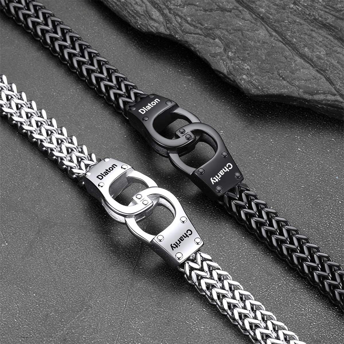 Two metal link bracelets with braided and chain-style links, one silver and one black, displayed diagonally on a dark textured surface with a slate prop. Both feature interlocking O-ring clasps with the brands 'Dion' and 'Charvy' engraved on the clasp plates.