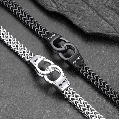 Two metal link bracelets with braided and chain-style links, one silver and one black, displayed diagonally on a dark textured surface with a slate prop. Both feature interlocking O-ring clasps with the brands 'Dion' and 'Charvy' engraved on the clasp plates.