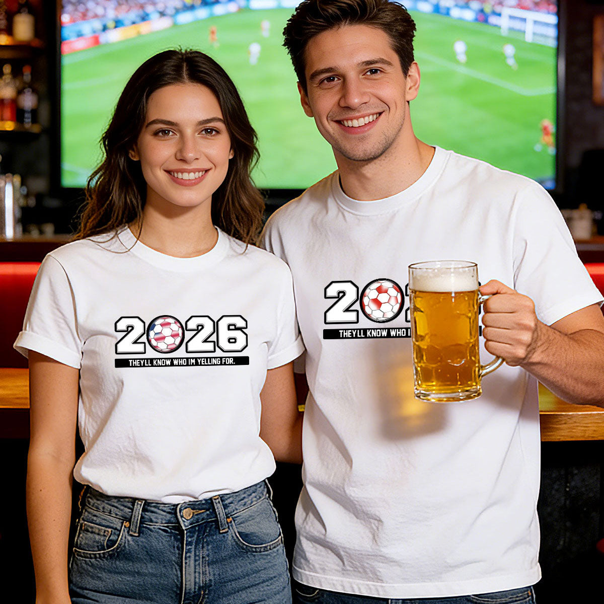 Two people wearing '2026' world cup t-shirts, one holding a beer mug, in front of a sports event on TV.