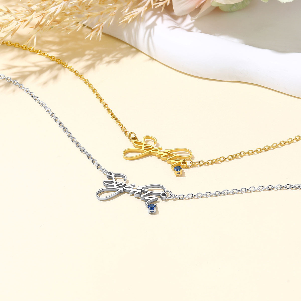Two personalized name necklaces with script lettering pendants, one in gold and one in silver, both with small blue stone accents, displayed on a light cream background with dried floral elements.