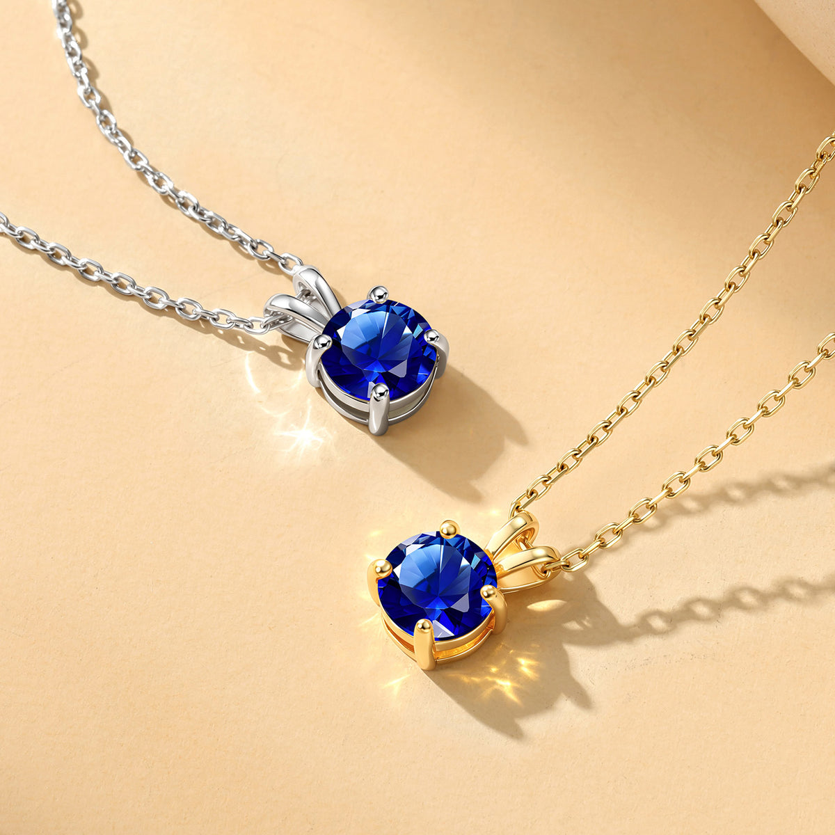Two round blue gemstone birthstone necklaces on delicate chains, one in silver-tone metal and one in gold-tone metal, displayed on a warm beige background.