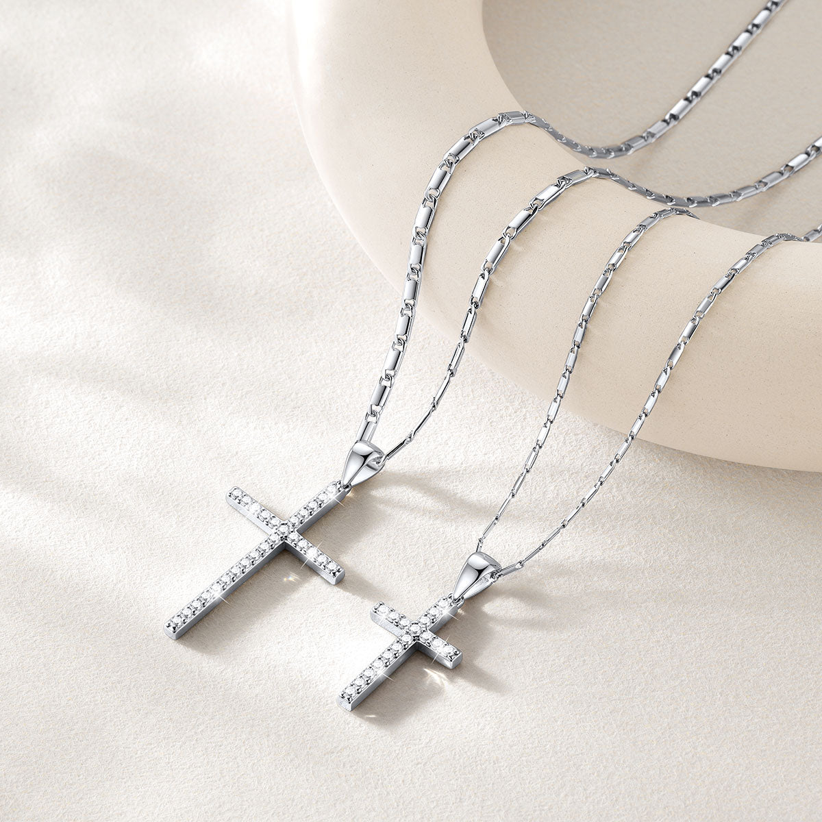 Two silver cross necklaces with pavé-set clear stones, displayed on a cream-colored curved stand against a soft beige background.