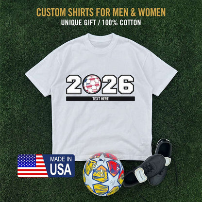 White t-shirt with '2026' world design on grass background, soccer ball, cleats, and USA made-in-USA badge.