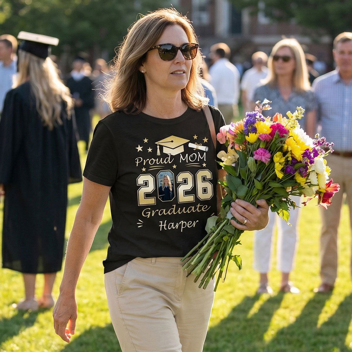 Woman in a 'Proud Mom 2026 Graduate' t-shirt holding flowers at a graduation ceremony.