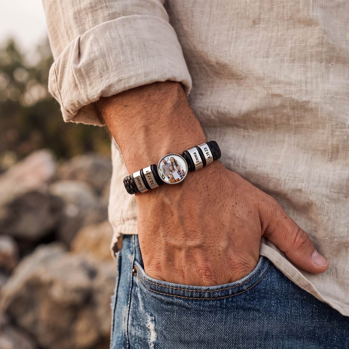 a man's wrist wearing a black braided leather photo bracelet with a custom circular dial photo featuring a couple, set against a blurred stone pavement background.