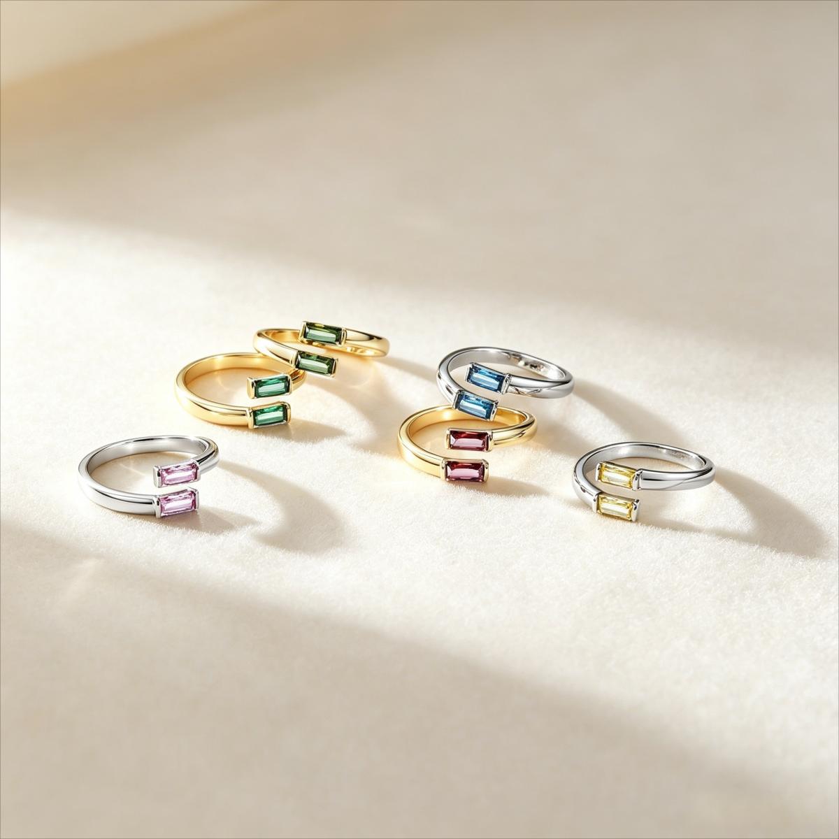 birthstone rings arranged on a warm cream surface, each featuring rectangular gemstones in different colors: green, blue, pink, yellow, and purple, with some rings in yellow gold and others in silver-tone metal.