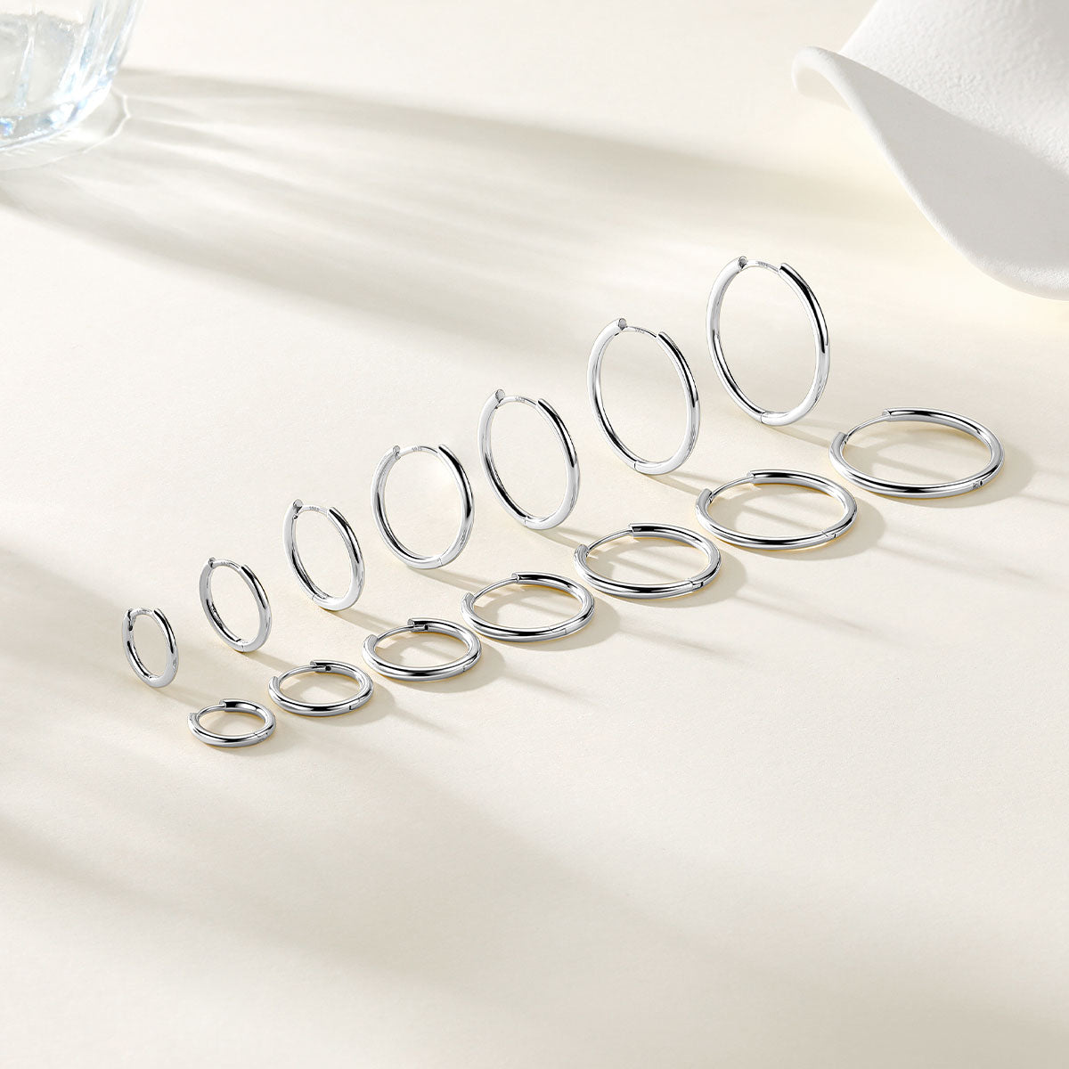 silver huggie earrings in multiple sizes, arranged diagonally on a light beige surface with soft shadows.